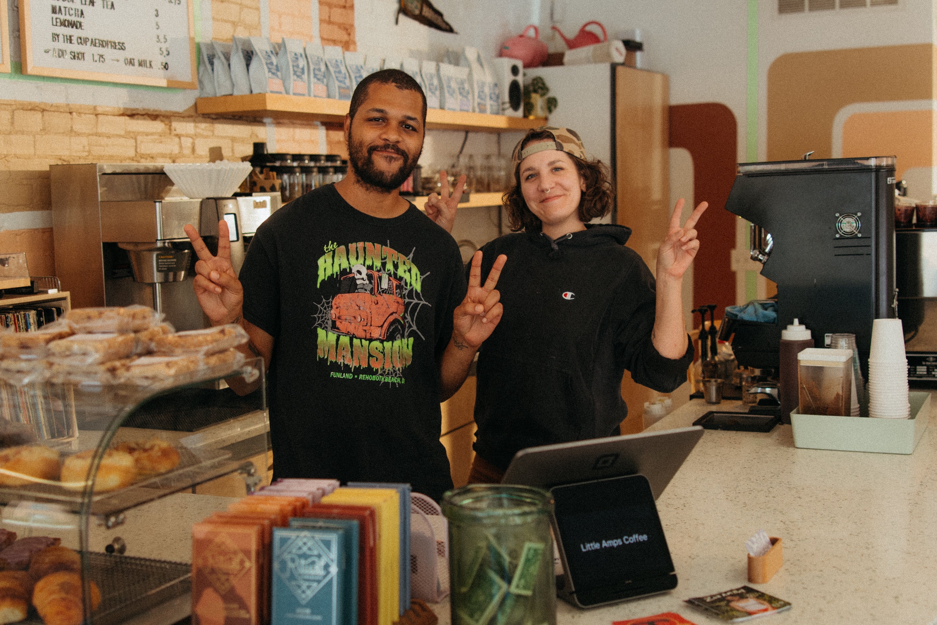 Two barista standing behind the counter of a coffee shop making peace signs surrounded by pastries, coffee beans, and an espresso machine.
