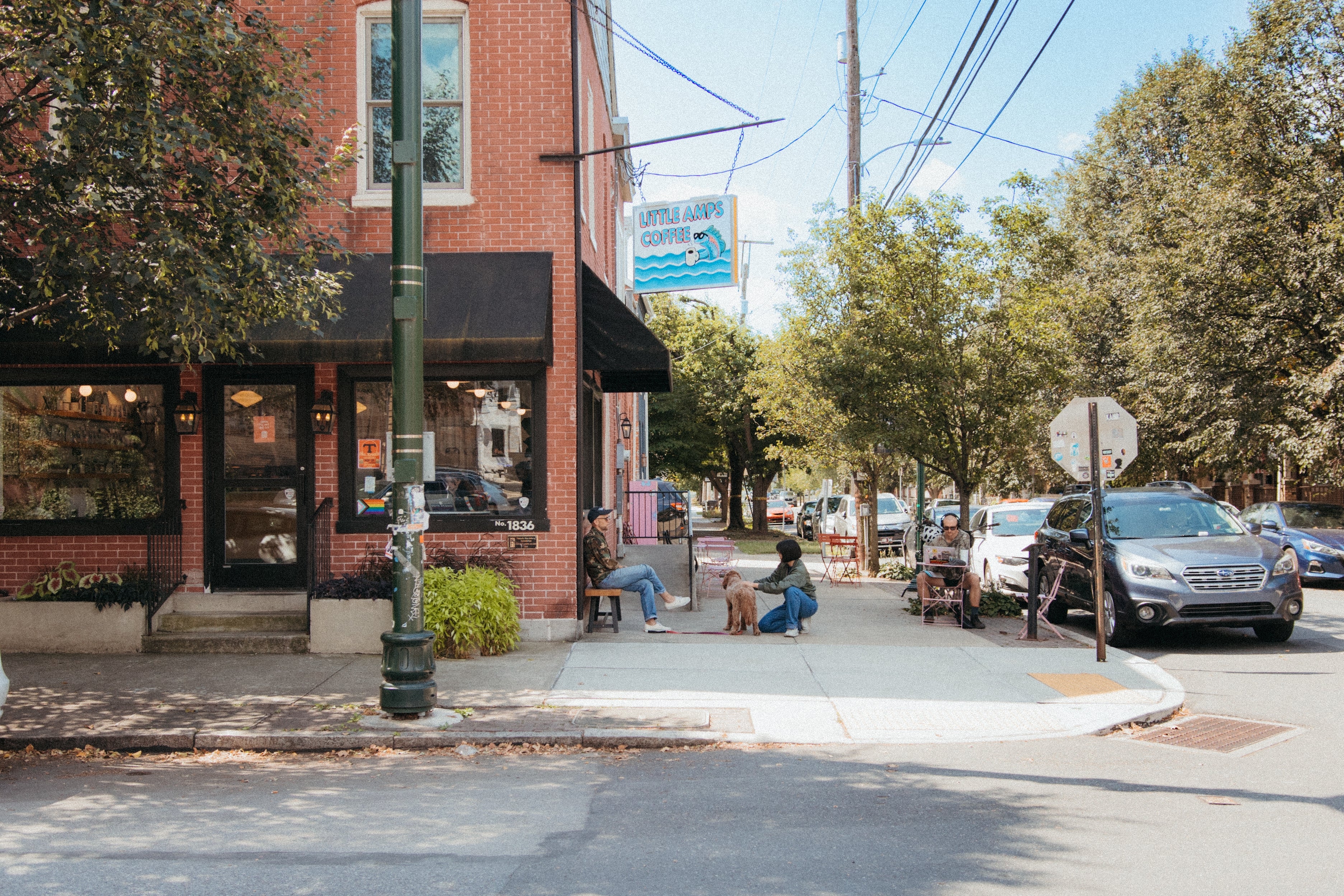 A coffee shop on the corner of a street with a man sitting outside on a bench while a woman pets a dog in front of him