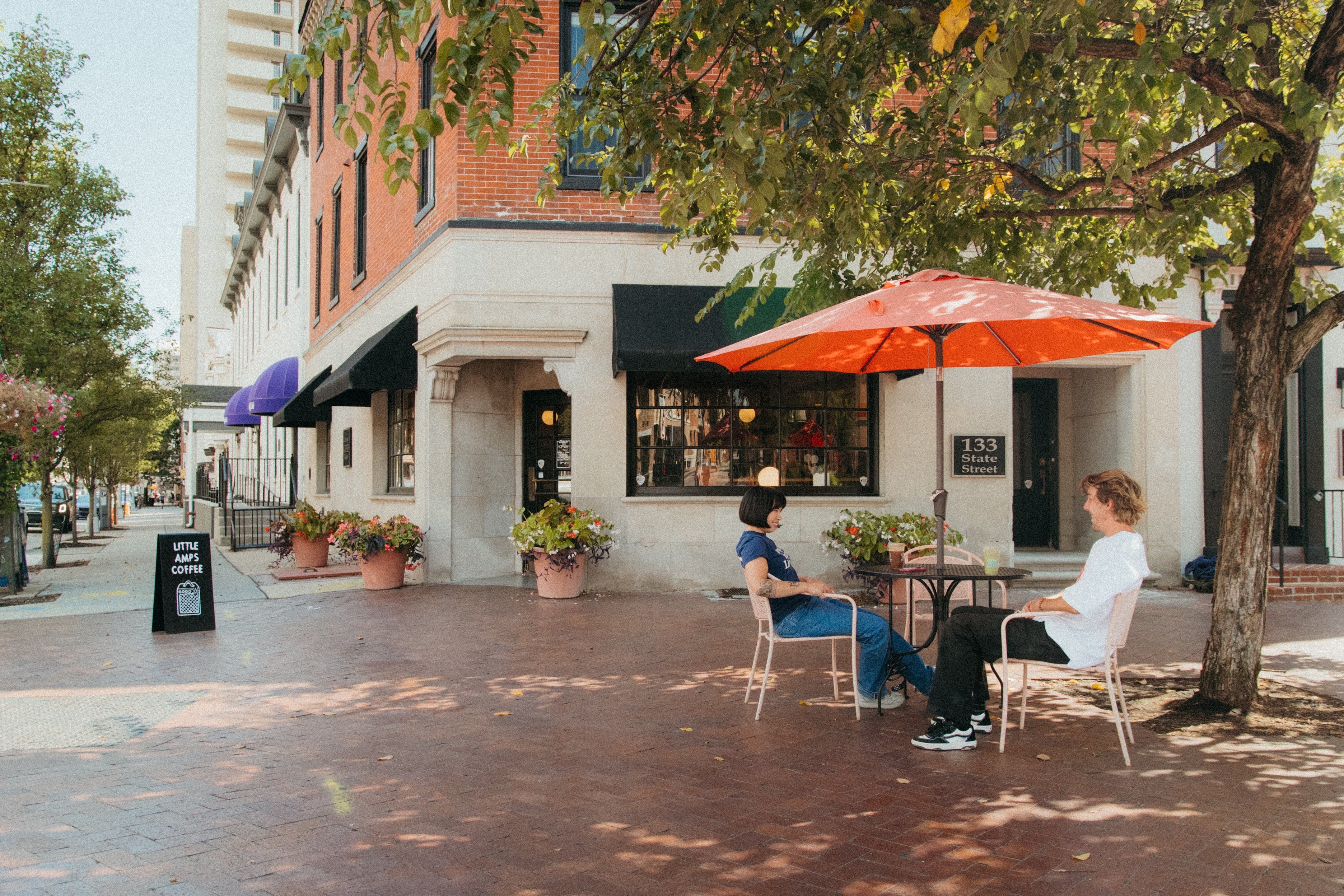 Two people sitting under an orange umbrella on a sidewalk in front of a coffee shop