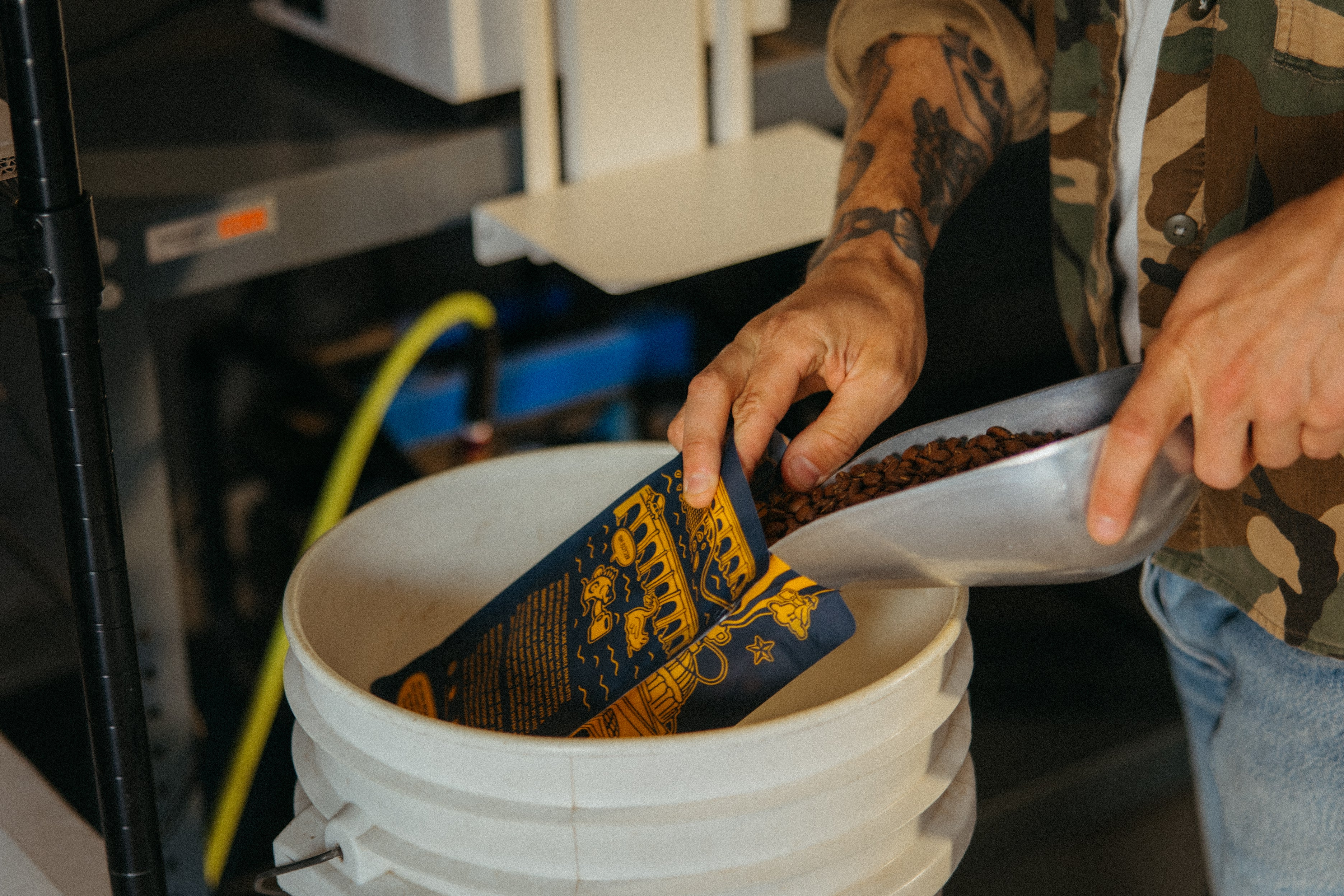 Person with tattoos scooping roasted coffee beans into bag of coffee.